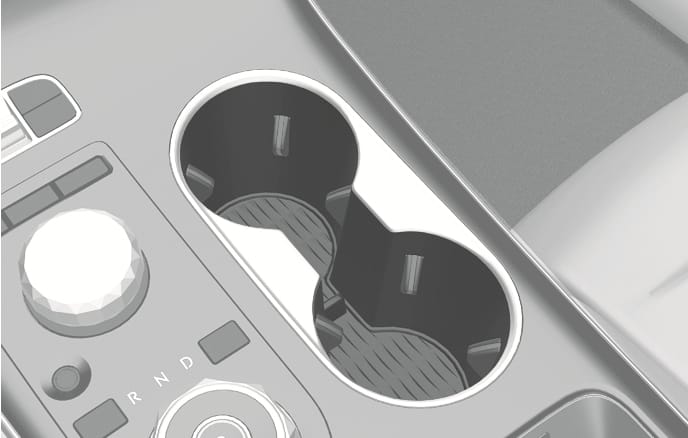 2025 Genesis GV70 front cup holder image. The image displays two circular cup holders located on the center console, positioned to the right of the rotary gear shift dial and controller. Internal support tabs are visible inside the holders. Explains the function of holding cups or small beverage containers securely.