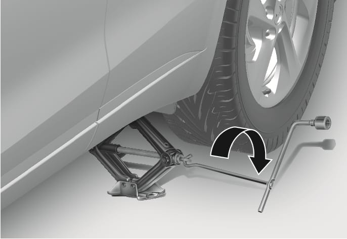 2025 Genesis GV70 jack operation image. It shows the jack handle inserted into the jack with the wheel lug wrench attached, and a black curved arrow indicating clockwise rotation. Explains the function of raising the vehicle.