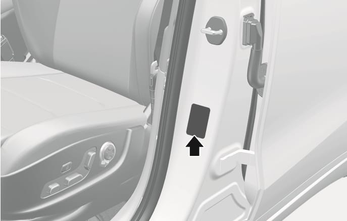 2025 Genesis GV70 tire specification and pressure label location image. A black arrow points upwards to the rectangular label attached to the driver's side center pillar. The driver's seat is visible on the left. Explains the location of the label providing recommended tire pressures.